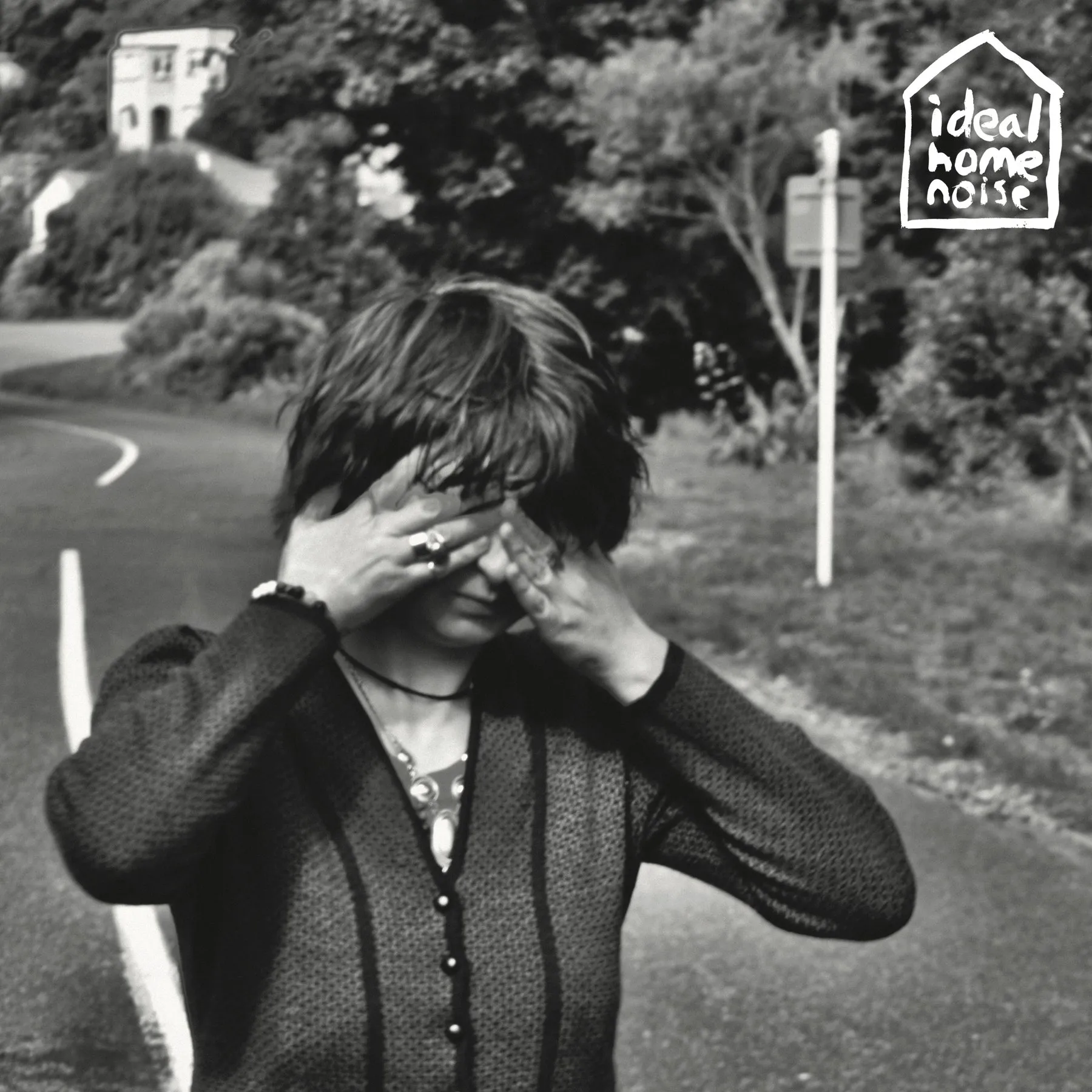 Cover for Ideal Home Noise — a black and white photograph of a figure standing on a road with both hands over their eyes, a house on the hillside behind, and a hand-drawn house-shaped mark in the top corner.