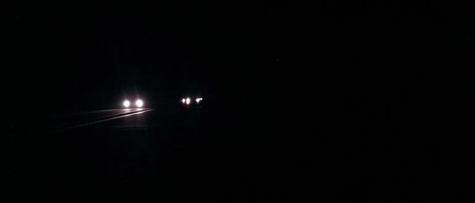 Headlights on a dark road, their beams fanning out over wet asphalt, another car's lights glowing faintly ahead.