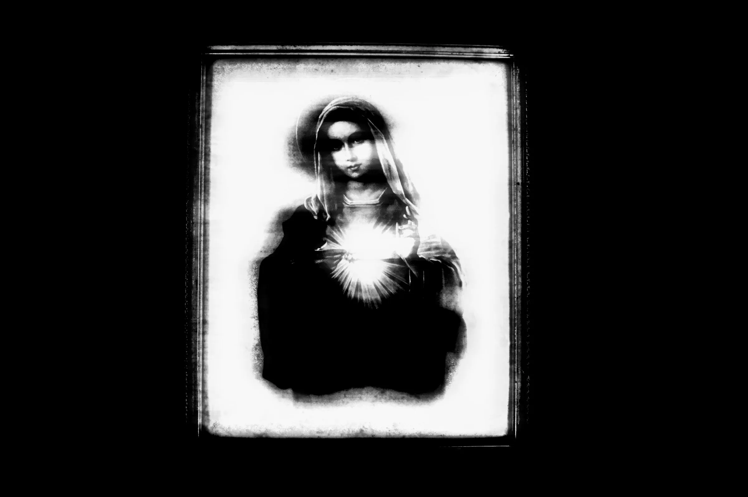 A framed Sacred Heart icon in stark black and white, the figure haloed, light bursting from the heart, the surrounding room entirely black.