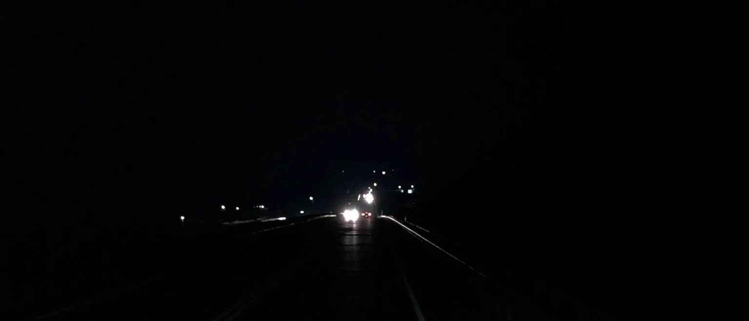A bridge at night, oncoming headlights and taillights glowing ahead, streetlights receding into the dark.